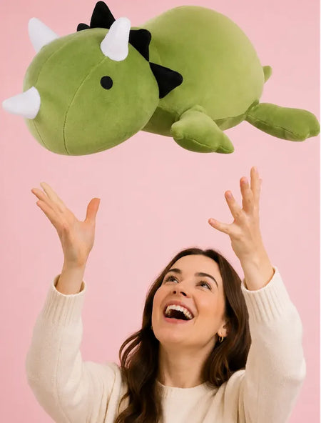 Woman tossing a green triceratops stuffed animal against a pink background